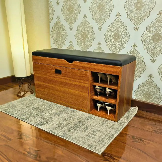 Wooden shoe rack cabinet with black cushioned top in a room with patterned wallpaper.
