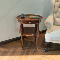 An oval wooden side table with a single drawer and lower shelf, styled with a blue cup and a book, positioned next to a cream-colored armchair in a room with wood flooring.