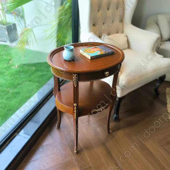 An oval wooden side table with a lower shelf, featuring gold-toned decorative legs and trim, styled with a small blue vase and a book in a bright room next to a white armchair.