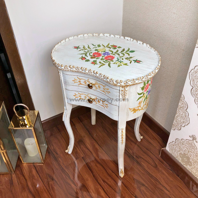 A wooden side table with a floral hand-painted design and metal trims, featuring a rectangular top and a pair of drawers on the base, placed in a room with an ornate wallpaper.