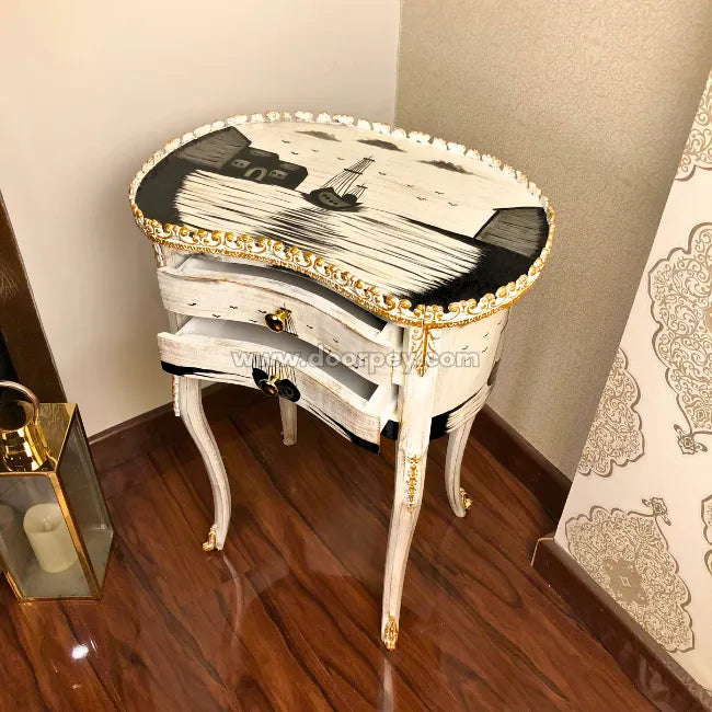 An ornate French-style wooden side table with a white and black painted design, gold trim, and carved legs, set in a room with a dark wood floor and patterned wallpaper.