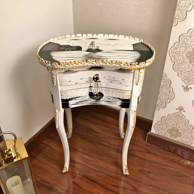 An ornate white and black hand painted wooden side table with gold trim, featuring a ship illustration on the top and decorative cabriole-style legs, shown in a home interior setting.