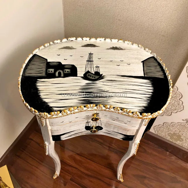 An ornate white and black hand painted wooden end table with gold trim, featuring a nautical scene with a ship on the tabletop and a small drawer with a gold-tone bell handle.