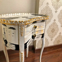 A decorative white and gold hand painted wooden table with a distressed finish, featuring hand-painted black ship and wave motifs on the front, gold ornate trim, and two gold handles, shown in an indoor room setting.