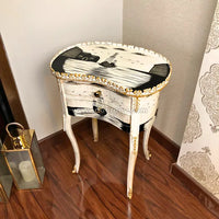 An ornate wooden table with a distressed white and black painted design, gold trim, and carved legs, placed on a dark wood floor in a room corner.