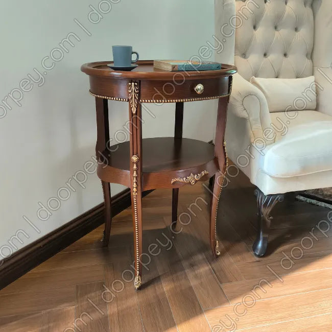 A round dark brown wooden side table with ornate gold-toned metal accents and a lower shelf, styled in a living room next to a tufted armchair on a wood floor.