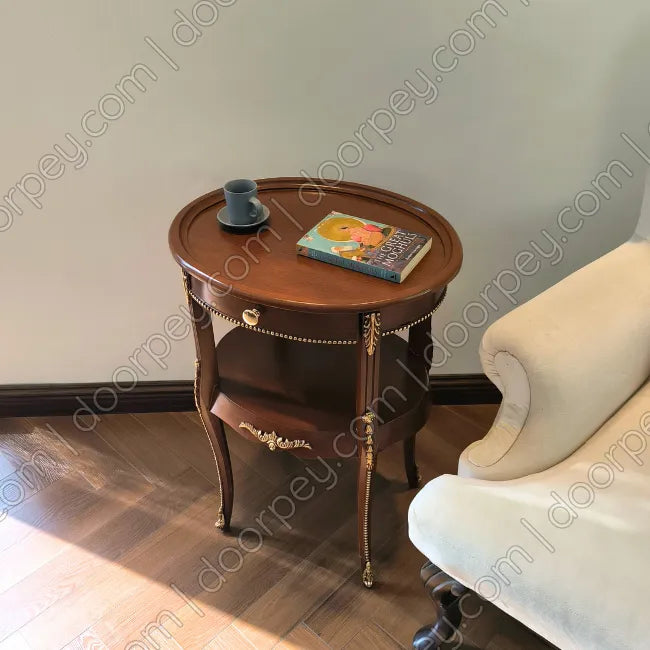 A walnut wooden side table with ornate gold-toned legs and trim, styled with a blue cup and a book, positioned next to a cream-colored armchair in a room with a light gray wall and wood floor.