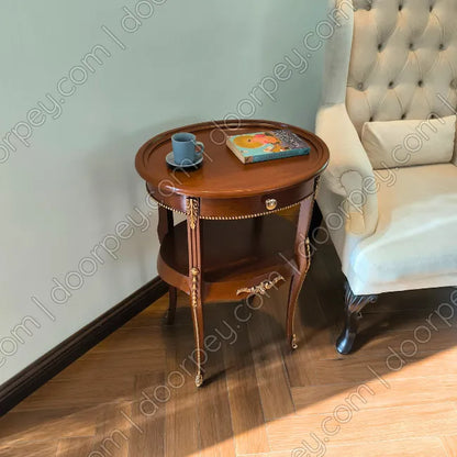 A round wooden side table with ornate gold-toned legs and trim, styled with a blue cup and a book, positioned next to a tufted armchair on a wood floor.