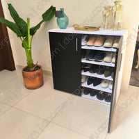 A modern white and black shoe cabinet with an open door showing multiple shelves of shoes, placed in a home entryway next to a potted plant. The shoe rack has decorative items on top, including vases and a tray.