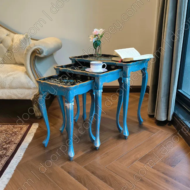 Three wooden nesting tables in a bright blue painted finish with black and gold hand painted patterned tops, styled in a living room corner next to a cream tufted armchair on a wooden floor.