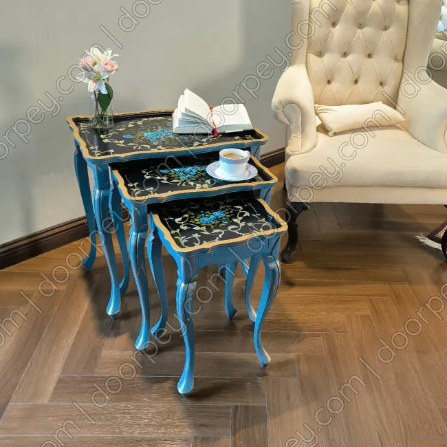 Three nested nesting tables with vibrant blue painted legs and black marble-patterned tops, styled with a vase of flowers, an open book, and a teacup on a wooden herringbone floor next to a cream tufted armchair.
