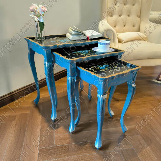 Three wooden nesting tables in bright turquoise blue with black and gold tops, styled in a living room setting with a tufted armchair and wooden floor.
