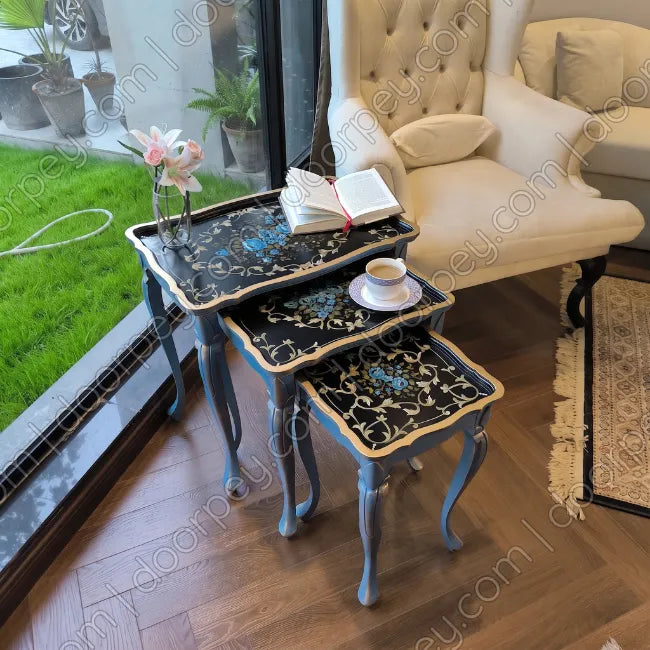 Three nested square nesting tables with ornate black floral-patterned tops and blue cabriole-style legs, styled in a bright room next to a window with a cream armchair and a view of green grass outside.
