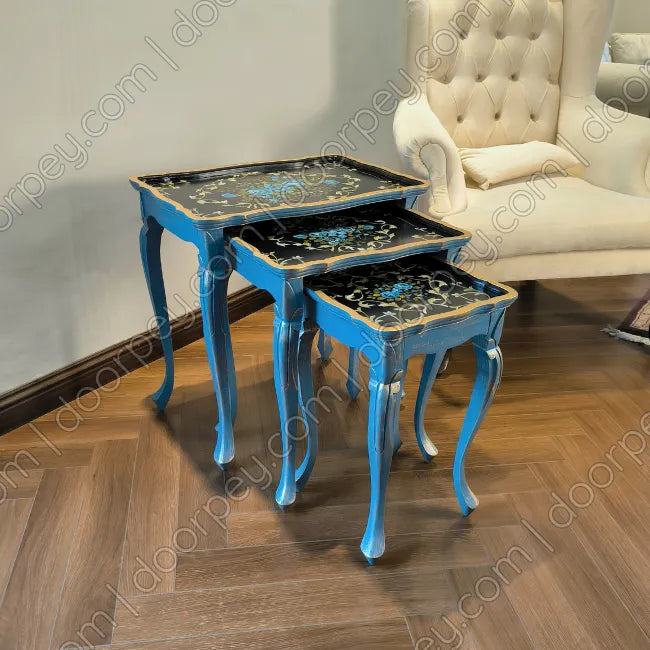 Three nested nesting side tables with curved blue legs and black mosaic-patterned tops, shown in a room setting with a wooden floor and a cream tufted armchair.