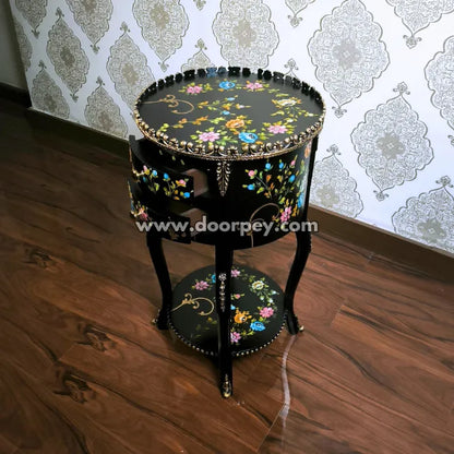 Decorative round wooden table with hand painted floral patterns and open drawers on a wooden floor and patterned wallpaper.