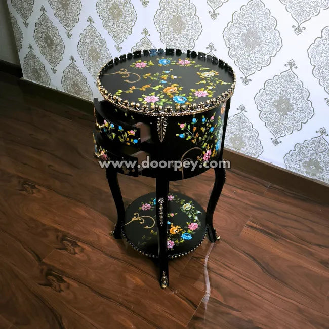 Decorative round wooden table with hand painted floral patterns and open drawers on a wooden floor and patterned wallpaper.