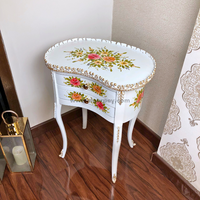 A white decorative wooden side table with painted patterns and gold trim on a wooden floor.