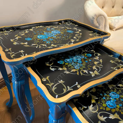 A set of three wooden nesting tables with black painted tops decorated with blue floral motifs and gold scrollwork, paired with blue legs, shown in an indoor setting.
