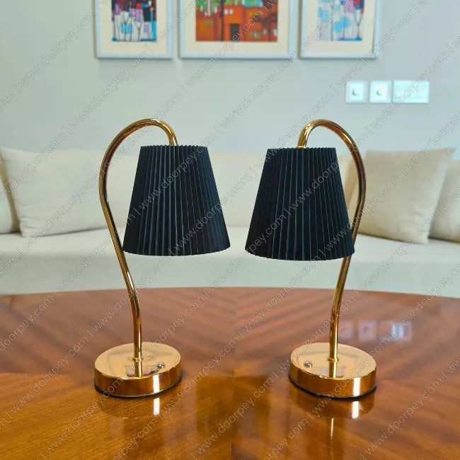 Two table lamps with black shades and gold bases on a wooden surface, with a sofa and framed pictures in the background.