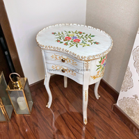 A wooden side table with a floral hand-painted design and metal trims, featuring a rectangular top and a pair of drawers on the base, placed in a room with an ornate wallpaper.