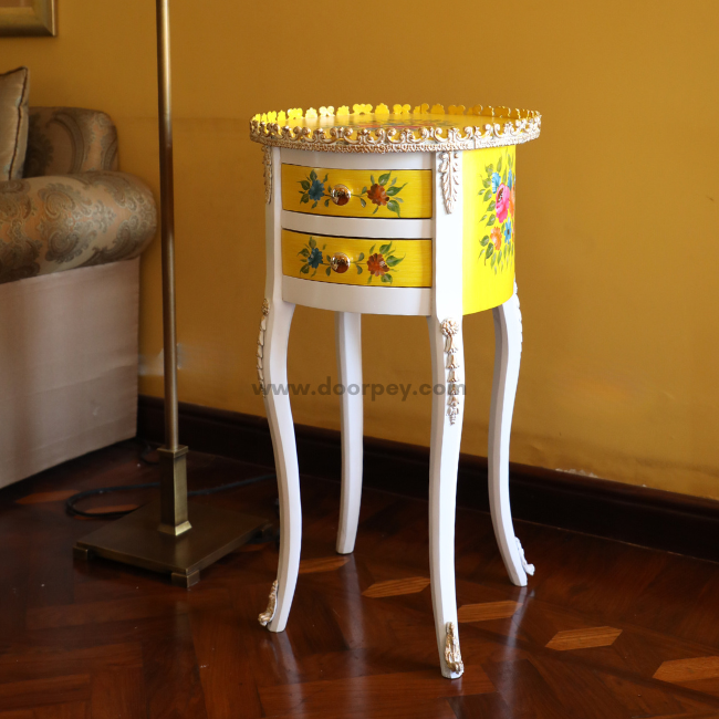 A round wooden side table with a yellow and white color scheme, adorned with metal trims and floral hand-painted designs. The side table is positioned in a room with a yellow wall and a white floor, next to a chair with a yellow upholstery.