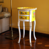 A round wooden side table with a yellow and white color scheme, adorned with metal trims and floral hand-painted designs. The side table is positioned in a room with a yellow wall and a white floor, next to a chair with a yellow upholstery.