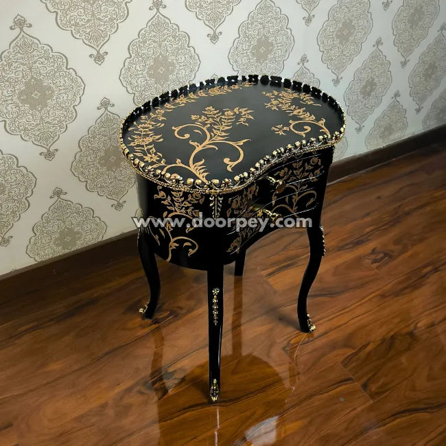 Decorative black end table with gold floral patterns on a wooden floor and patterned wallpaper.
