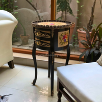 A round wooden table with metal trims, ornate design, and a floral pattern on the drawer front, placed in a room with a plant and seating in the background.