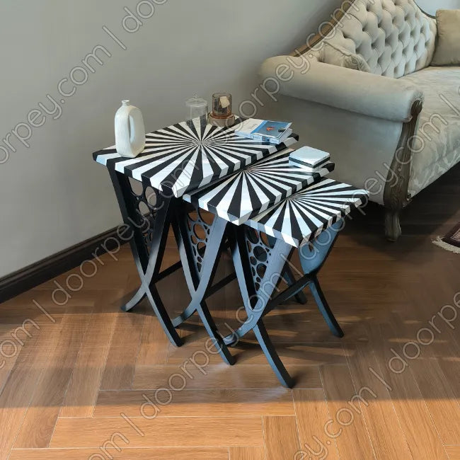 Three nested nesting tables with black and white striped starburst-patterned tops and dark wooden legs, styled in a living room next to a beige sofa on a wooden floor.