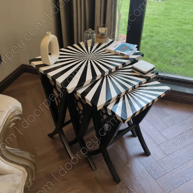 A set of three wooden nesting side tables with black and white striped starburst-patterned tops and black legs, styled in a room with a window overlooking a green lawn.