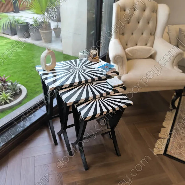 Set of three nesting side tables with black and white striped starburst-patterned tops and black frames, styled in a modern living room next to a tufted armchair.