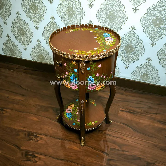 Round wooden end table with floral patterns on a wooden floor and patterned wall background.