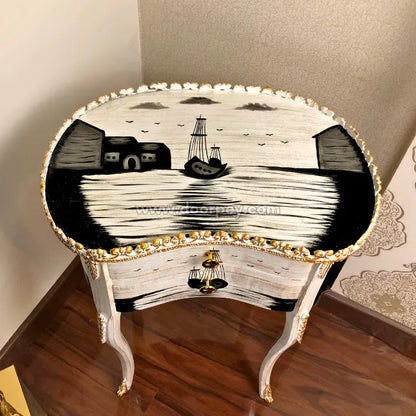 An ornate white and black hand painted wooden end table with gold trim, featuring a nautical scene with a ship on the tabletop and a small drawer with a gold-tone bell handle.